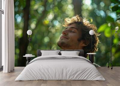 A young man with curly hair breathes deeply in a lush green forest on a sunny day Wall mural