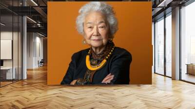 An elderly Asian woman with white hair and a serious expression poses for a portrait. She is wearing a black jacket, a yellow necklace, and gold earrings. The background is a solid orange color Wall mural