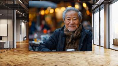 An older man with a beard and gray hair is smiling at the camera. He is wearing a black coat and a scarf. The scene takes place in a restaurant with tables and chairs Wall mural