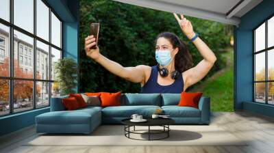 A young caucasian woman wearing a mask with headphones and smart phone taking selfie Wall mural