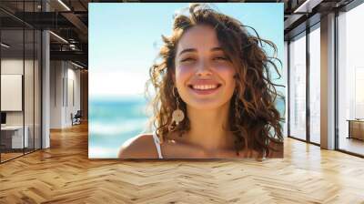 Smiling Young woman with curly hair standing on a sandy beach with the ocean in the background. Wall mural