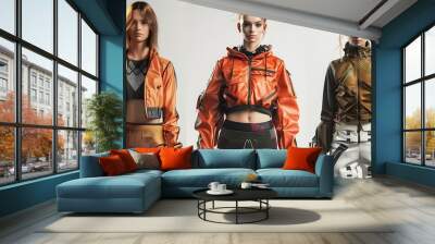 Three young women in futuristic stylish clothing posing against a white background. They are all wearing jackets, pants, and other accessories. Wall mural