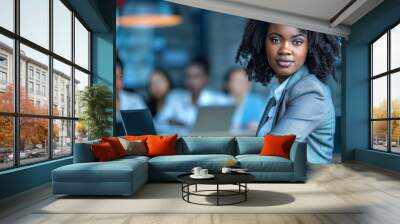 Attractive african young confident businesswoman sitting at the office table with group of colleagues in the background, working on laptop computer - generative ai Wall mural