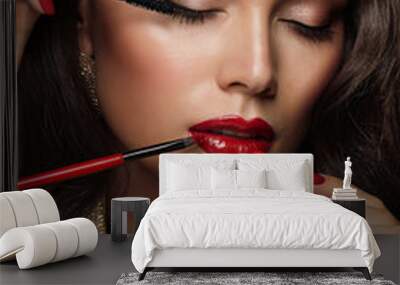 Woman applying Make up. Beauty Model Girl put on Red Lipstick. Professional Makeup Artist painting Lips, holding Mascara and Eye Shadow Brush. Elegant Lady Portrait with closed Eyes Wall mural