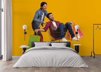 Two young men laughing with one of them sitting in a trolley holding carry bags.	 Wall mural