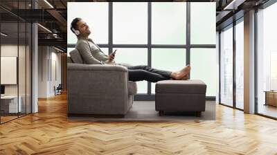 Young businessman listening music on headphone using mobile phone while leaning on sofa at home Wall mural