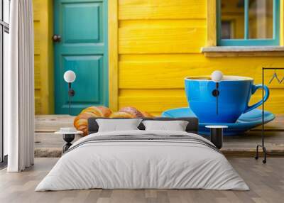 A blue coffee cup and saucer sit on a rustic wooden table, accompanied by two golden croissants, creating a delightful breakfast scene against a backdrop of a yellow and blue wooden exterior. Wall mural