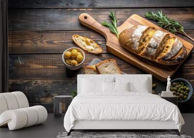 Rustic wooden table with fresh baked bread, sliced and ready to be enjoyed, accompanied by a bowl of olives and sprigs of rosemary. Wall mural