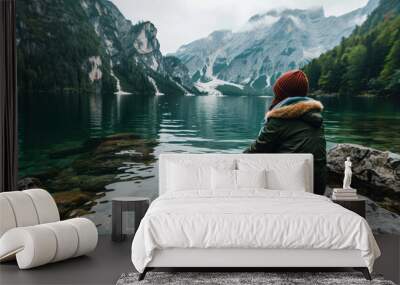 Back view of a girl traveler sitting on a stone shore by a lake against the background of mountains in a warm hat and autumn jacket. Travel time. Wall mural