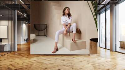 Beautiful woman fashion model brunette hair tanned skin wear white overalls button suit sandals high heels accessory bag clothes style journey safari summer collection plant flowerpot wall stairs. Wall mural