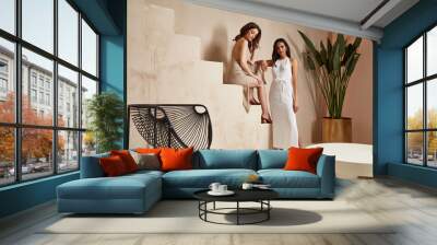 Two sexy beautiful woman friends brunette tanned skin makeup cosmetic fashion clothes summer collection cotton dress style summer journey walk date beach wear interior stairs leaves flowerpot. Wall mural