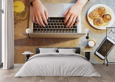 Girls hands are typing on  laptop on grey grunge kitchen table with phone, candlestick, mug and plate with buns on it. Working and studying from home concept.
 Wall mural