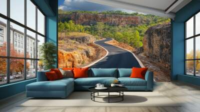 A winding asphalt road leads through a scenic mountainous landscape with lush greenery, rocky cliffs, and a blue sky with clouds. Wall mural