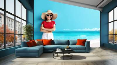 Smiling woman in a red top and white shorts, wearing a sunhat and sunglasses, standing on a sunny beach with a blue sky and ocean backdrop. Wall mural