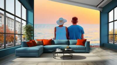 Two people sitting on the beach during a beautiful sunset, enjoying the serene coastal view and calm ocean under a vibrant sky. Wall mural