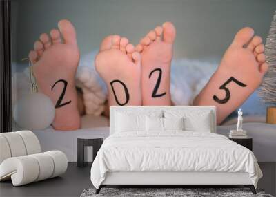 Close-up of two children's bare feet sticking out from under white blanket on bed with 2025 written on it. Winter New Year concept. Place for text. Wall mural