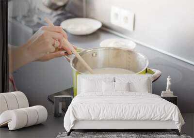 Woman preparing bechamel sauce or cream in a pan. Woman hand is mixing boiling milk with wooden spoon. Wall mural