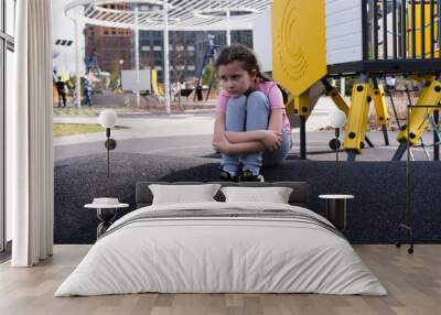 Girl alone with no one to play with. Sad sad child sits against the backdrop of bright playground where other children are playing and having fun. Concept of longing, disappointment, grief and despair Wall mural