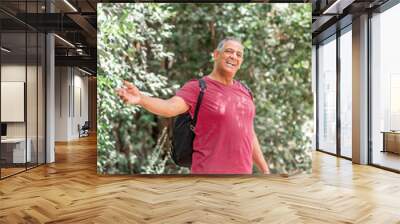 Portrait of a happy activity athlete senior man carrying a backpack, enjoying beautiful view. Healthy old man on a hiking trip. Male tour guide in the forest doing a hand inviting gesture. Wall mural