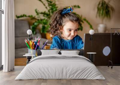 Happy little girl in headphones smiling and making notes in notepad while communicating with teacher through video chat app during online education at hom Wall mural