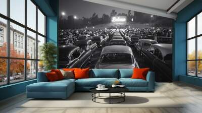 Under the night sky, vintage cars fill a drive-in theater lot, illuminating the surroundings. A large neon sign glows brightly, attracting moviegoers to enjoy a film together Wall mural