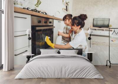 A little girl and her mother are cleaning the kitchen. a woman and a child wipe the oven in the kitchen. house cleaning. helping mom. Wall mural