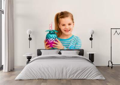 Little blonde girl is smiling on a white background. child girl in a blue jacket is holding a pineapple-shaped drink glass on a white background. summer vacation concept Wall mural