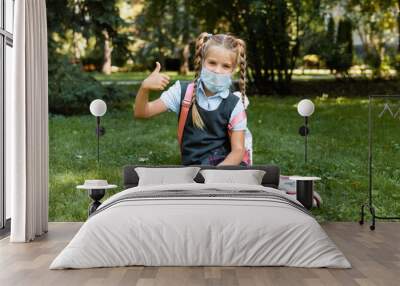 little schoolgirl in a medical mask sits in the park on the grass and has fun. Wall mural