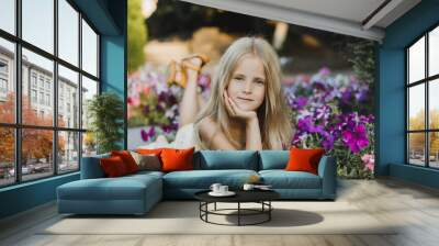 portrait of a cute girl 5-6 years old with a petunia flower, sitting over flowering bushes in the garden outdoors. Spring season. Stylish little child against the backdrop of nature. Spring. Wall mural