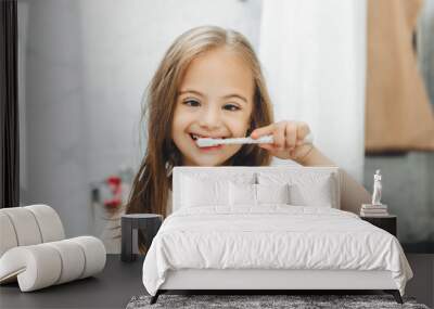 Portrait of a girl with Down syndrome brushing her teeth. Happy little girl brushing her teeth in front of a bathroom mirror. Morning hygiene. Wall mural