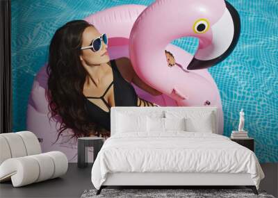 Hot and fashionable brunette model girl with perfect sexy body in stylish black bikini and glamorous sunglasses, sits on the floating pink flamingo and posing at the swimming pool Wall mural