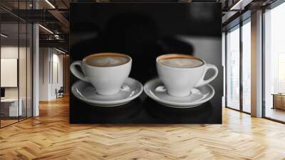 Two white cups of Cappuccino coffee with heart-shaped milk foam. Side view on two cups of latte coffee with heart figure on milk foam on black background. Lifestyle Wall mural