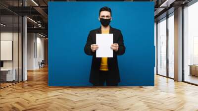 Young man in a black coat and black protective mask posing with an empty sheet of paper at the blue background, isolated with copy space. Promotion concept. Healthcare concept Wall mural