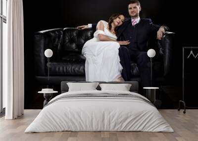 Engaged couple wearing modern wedding suit and bridal dress in a classical retro fashion style.  The stylish groom and bride are sitting on a leather couch in a studio. Wall mural