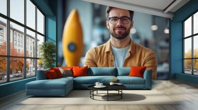 A bearded man with glasses, wearing a brown shirt, smiles confidently in an office environment with a blurred rocket model in the background Wall mural