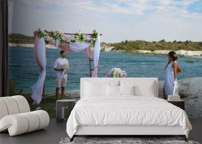 the groom in a white shirt with a bouquet of white flowers stands near the blue lake Wall mural