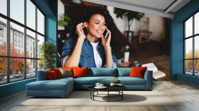 African-american young woman talking on phone in her cosy house. Businesswoman working from home office and using cell phone. Beautiful afro american woman sitting at the desk in a home office. Wall mural