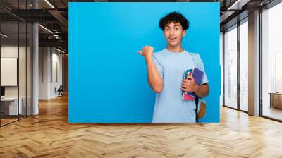 Amazed boy guy high school male pupil student teenager with bag holding books copybooks pointing finger empty copy space for advertisement isolated on blue background. Education back to school concept Wall mural