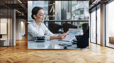 Asian businesswoman holding a paper in hand and showing analitics while talking to her business colleague Wall mural