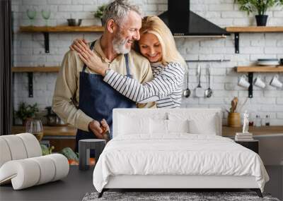 Beautiful mature couple hugging while cooking vegetable salad at kitchen table Wall mural