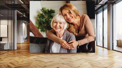 Cheerful middle aged woman embracing senior mother at home and looking at camera Wall mural