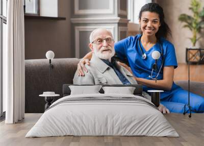 Front view portrait of caring afro nurse taking care of an elderly caucasian man grandfather grandpa indoors. Happy female doctor consulting old man in clinic. Wall mural