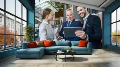 Group of coworkers in suits working together on digital tablet. Teamwork and collaboration in a modern workplace office. Business partners planning a project looking on pad computer Wall mural