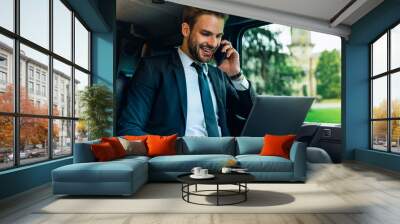 Handsome young man in full suit talking on smart phone and working on laptop, discussing job questions and smiling while sitting in the car Wall mural