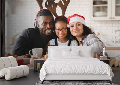 Happy african-american family, parents and small daughter in Santa Claus hat celebrating Christmas and New Year together at home, looking at camera during holiday breakfast with presents gifts Wall mural