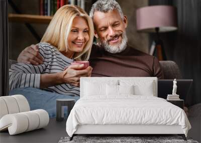 Happy beautiful middle aged couple using laptop and smiling while resting on couch at home Wall mural