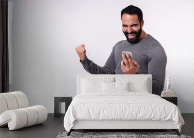 Happy excited young Arabian man in casual attire using mobile phone isolated over white background Wall mural