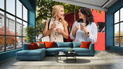 Happy smiling caucasian women sisters best friends girlfriends communicating talking spending time together while drinking hot beverage coffee from paper cups outdoors. Wall mural