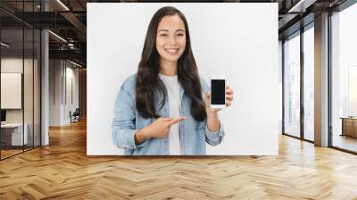 Happy young asian woman holding blank screen mobile phone and pointing finger over white background Wall mural