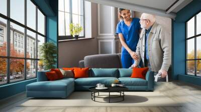 Portrait of an african young nurse helping old elderly disable man grandfather to walk using walker equipment in the bedroom. Senior patient of nursing home moving with walking frame and nurse support Wall mural